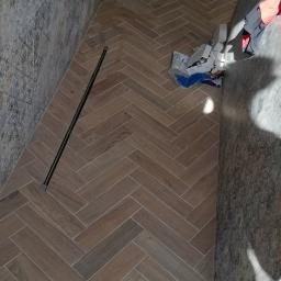 Mxbud - Ułożona w jodełkę podłoga z płytek imitujących drewno w kabinie prysznicowej we Wrocławiu, z widocznym odpływem liniowym i fragmentem ściany wyłożonej płytkami o wzorze przypominającym kamień.
