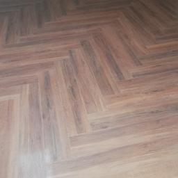 Mxbud - Ułożona pod kątem brązowa podłoga z płytek imitujących drewno, widoczne łączenia fugi i fragment białej ściany przy podłodze.