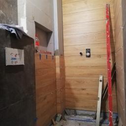 Mxbud - Wnętrze łazienki w trakcie układania płytek imitujących drewno na ścianach, widoczny poziomica, krzyżyki dystansowe i instalacja podtynkowa WC, w tle surowy beton i butelka napoju.