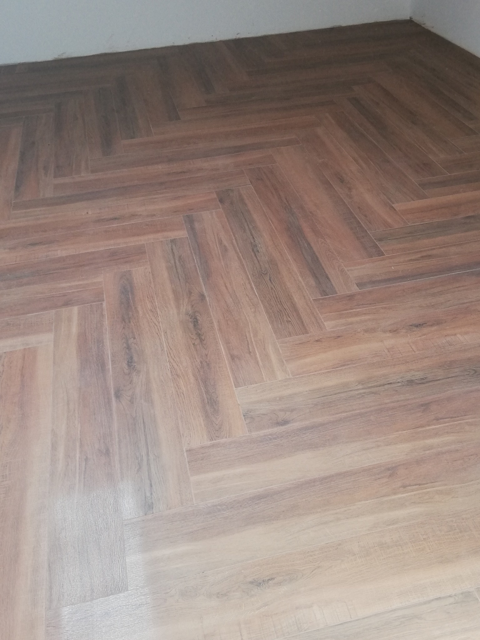 Ułożona pod kątem brązowa podłoga z płytek imitujących drewno, widoczne łączenia fugi i fragment białej ściany przy podłodze.