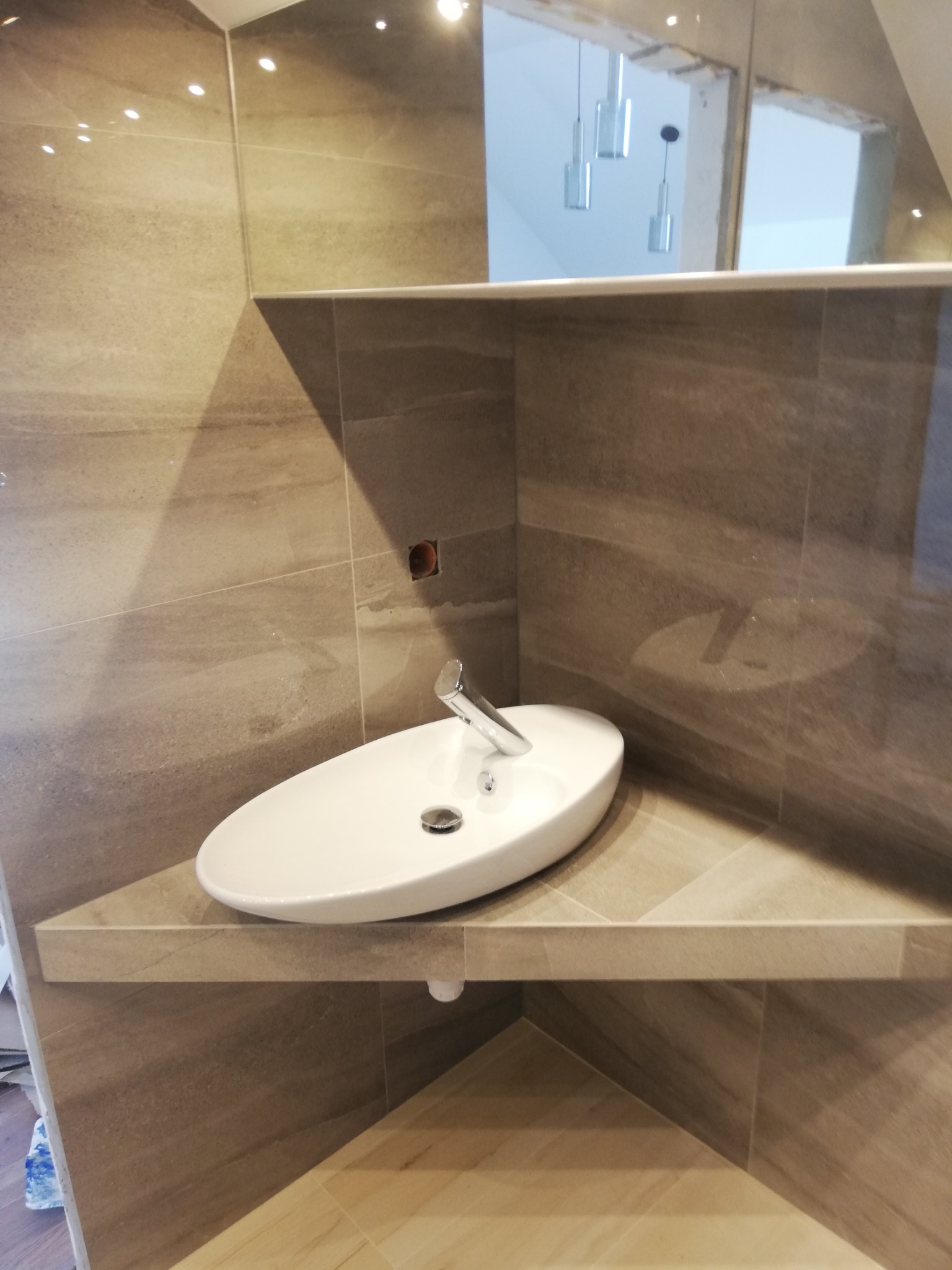 Nowoczesna, owalna umywalka nablatowa na półce z płytek w narożniku łazienki, z widocznym fragmentem lustra i minimalistycznymi lampami w tle.