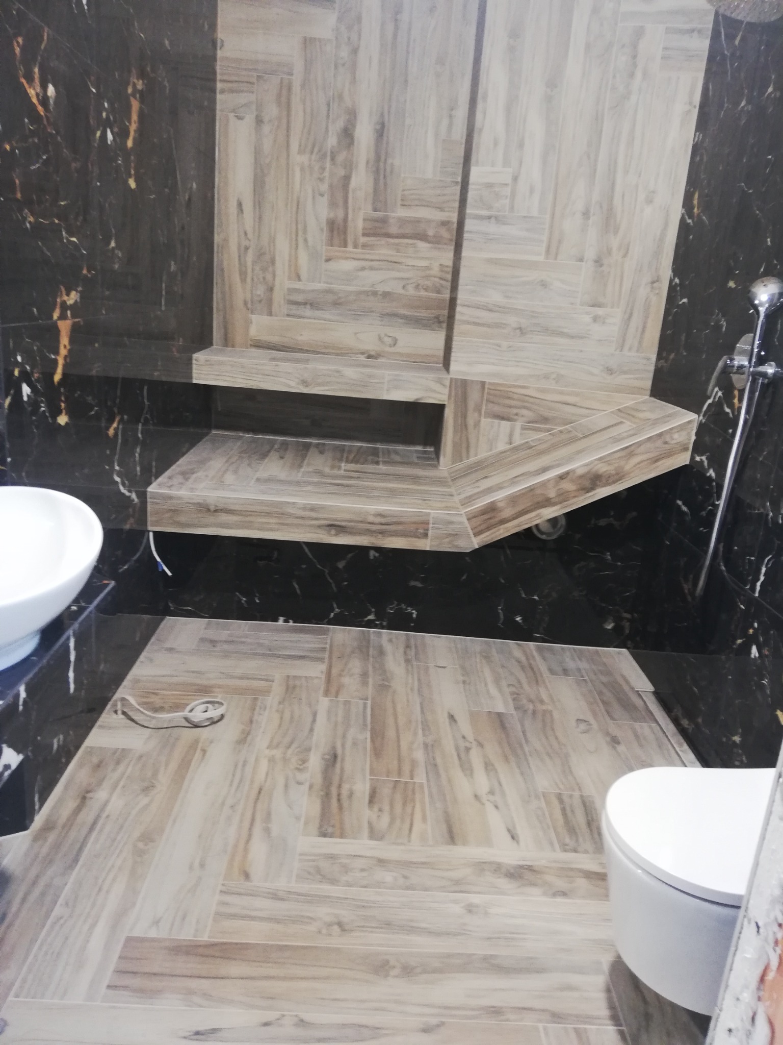 Wykończona łazienka z czarnymi marmurowymi ścianami i podłogą oraz prysznicem wyłożonym płytkami imitującymi drewno ułożonymi w jodełkę, widoczna umywalka i toaleta.