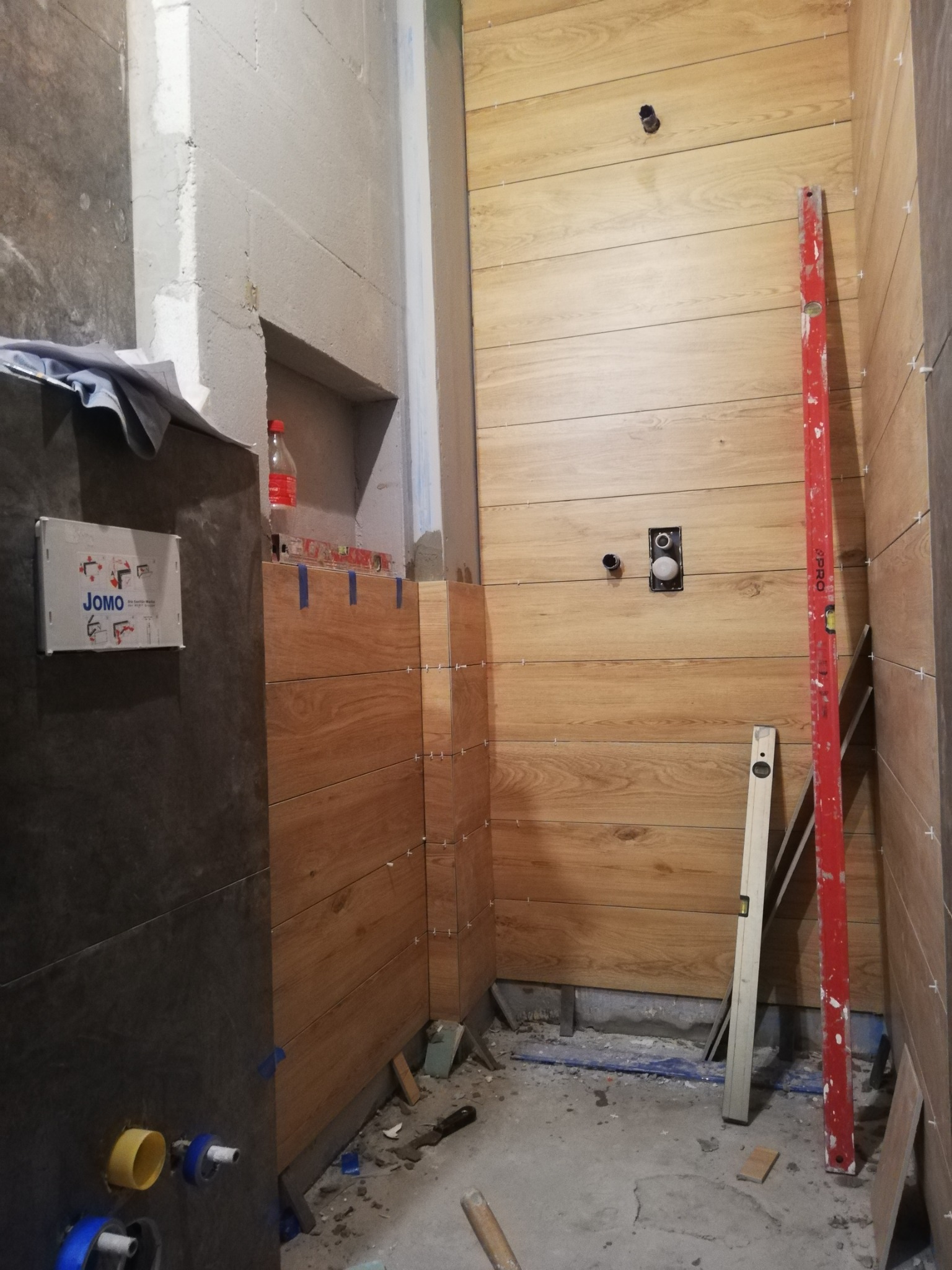 Wnętrze łazienki w trakcie układania płytek imitujących drewno na ścianach, widoczny poziomica, krzyżyki dystansowe i instalacja podtynkowa WC, w tle surowy beton i butelka napoju.