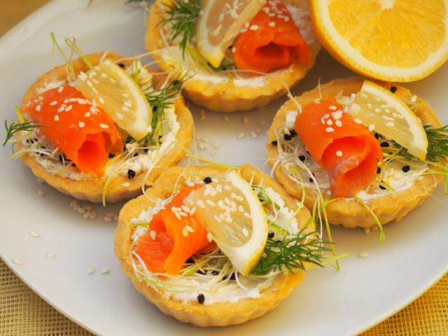 Cztery tartaletki z kremowym serem, łososiem rolowanym, plasterkiem cytryny, koperkiem i posypane sezamem, ułożone na białym talerzu obok połówki cytryny.