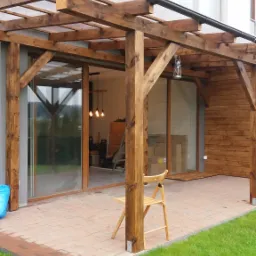 Drewniana pergola z przezroczystym zadaszeniem przylegająca do budynku z drewnianą elewacją, widok na taras z kostki i trawnik, krzesło pod pergolą.