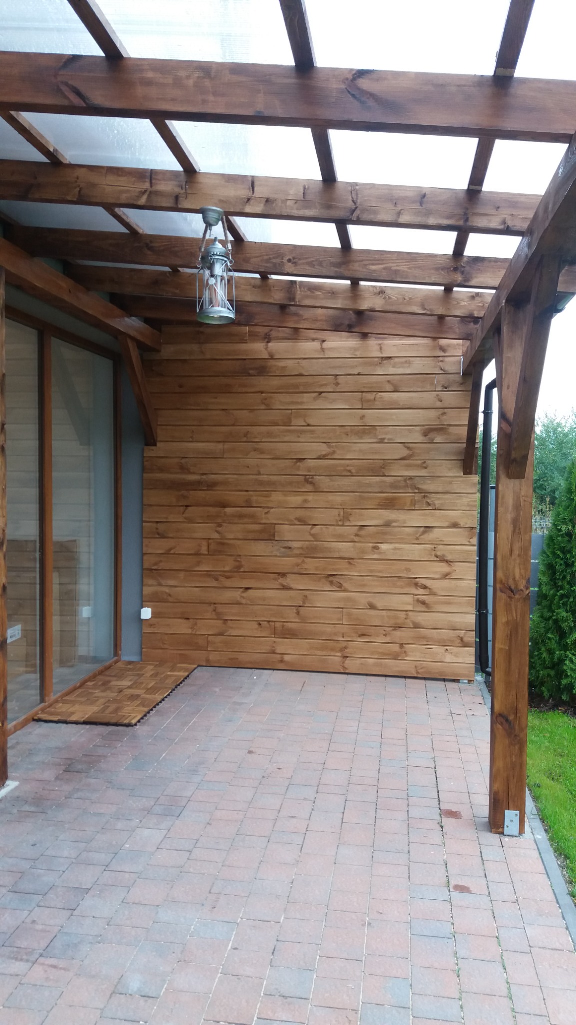 Zadaszony taras z drewnianą konstrukcją i ścianą z desek, oświetlony wiszącą latarnią, wyłożony kostką brukową.