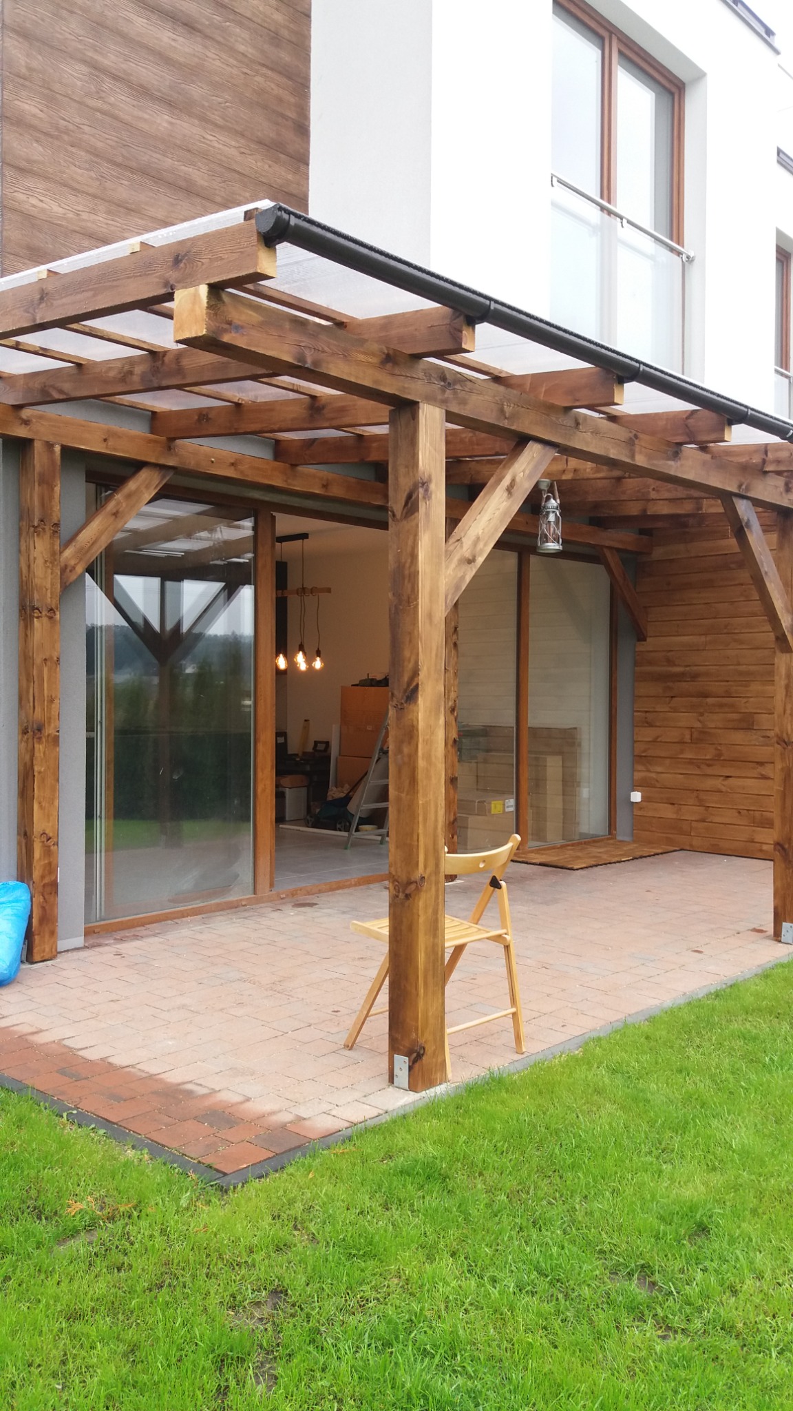 Drewniana pergola z przezroczystym zadaszeniem przylegająca do budynku z drewnianą elewacją, widok na taras z kostki i trawnik, krzesło pod pergolą.