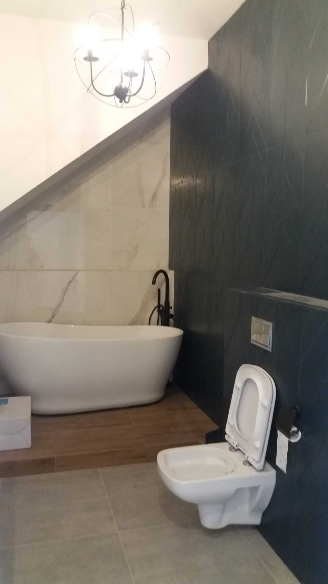 Nowoczesna łazienka na poddaszu z wolnostojącą wanną, czarną armaturą i toaletą podwieszaną na tle ściany z ciemnymi płytkami o geometrycznym wzorze.