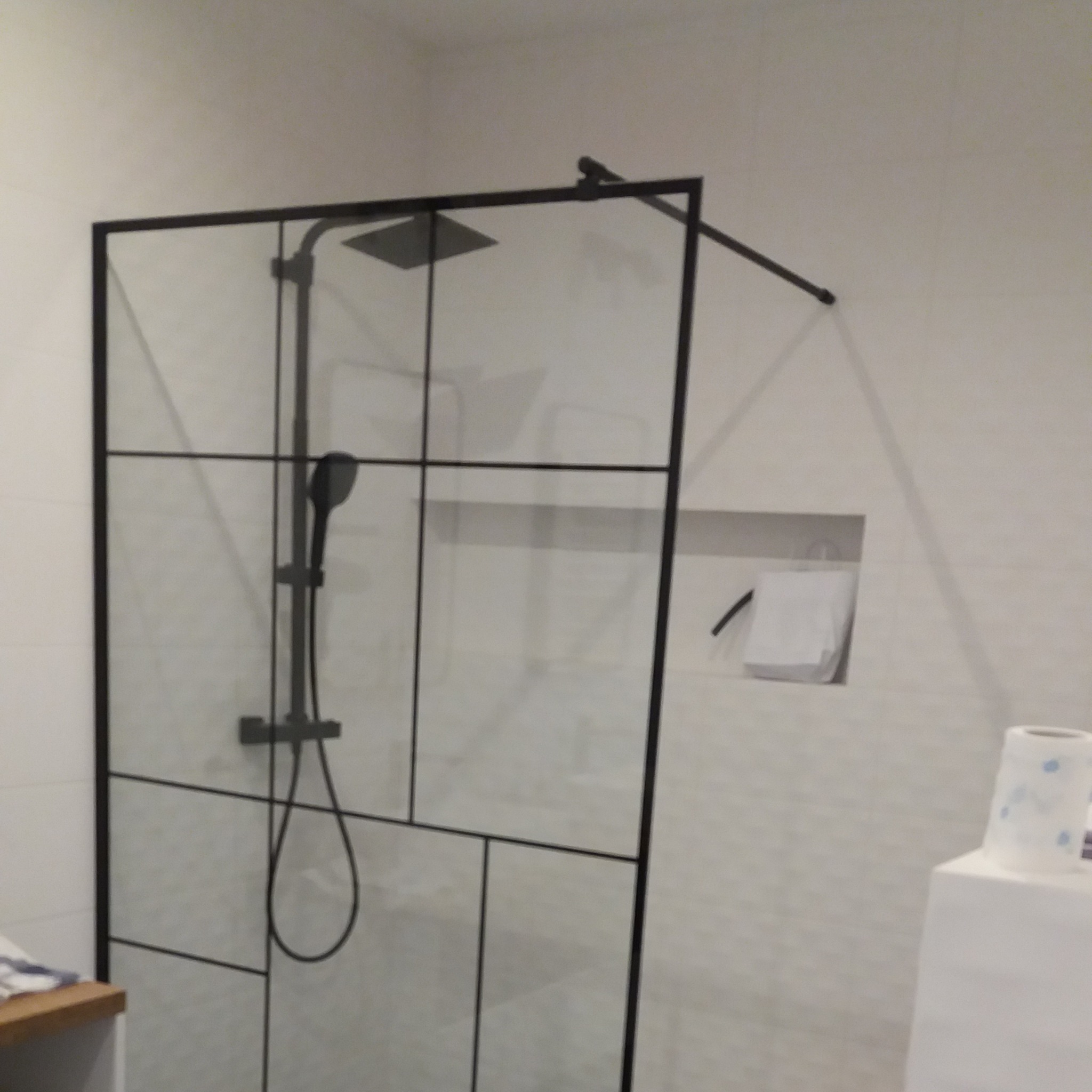 Kabina prysznicowa z czarnymi profilami w nowoczesnej łazience z wnęką na kosmetyki i widocznym dozownikiem mydła w tle.