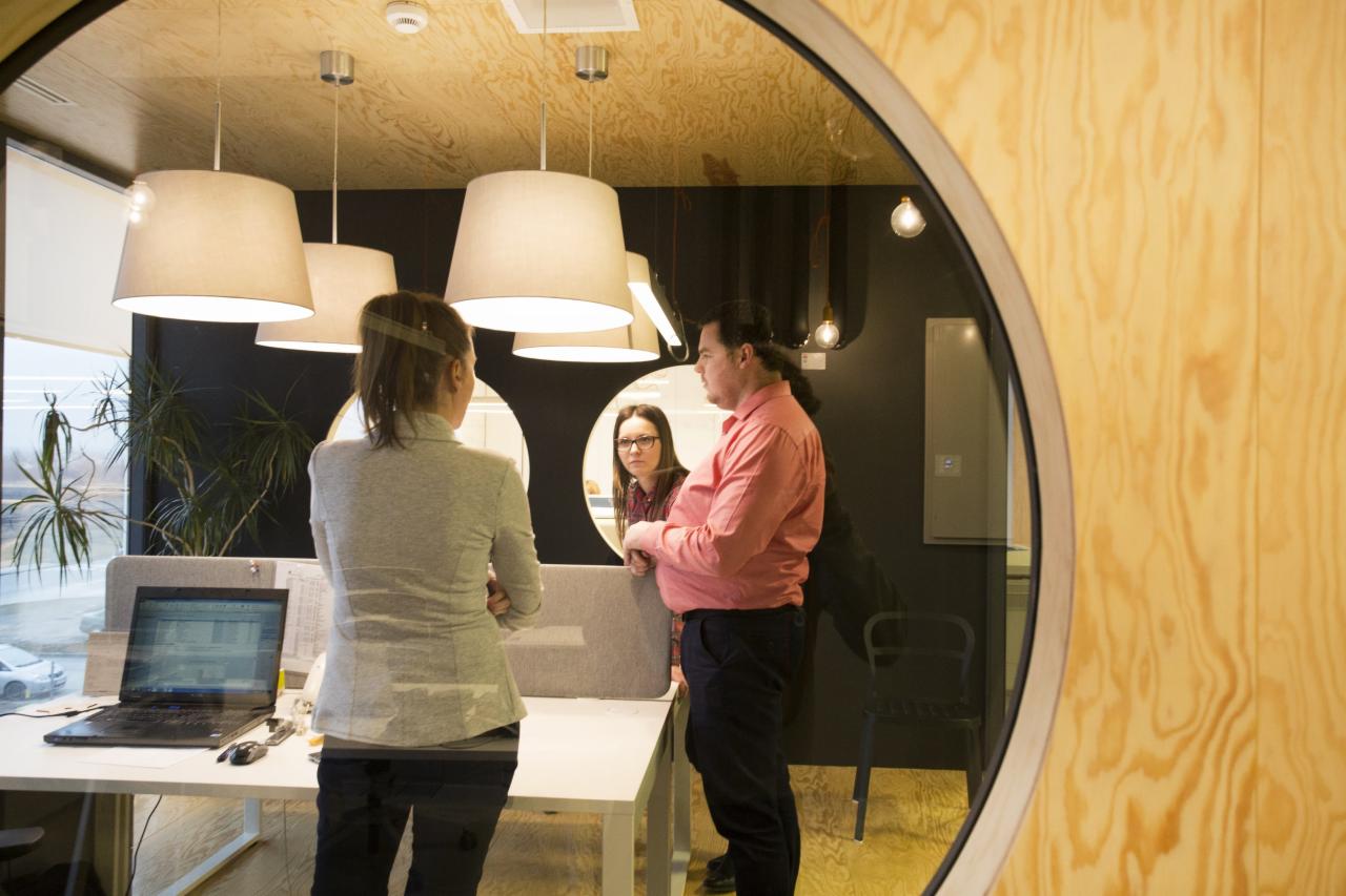 Wnętrze biura z trzema osobami w trakcie rozmowy widoczne przez okrągłe okno, w tle wiszące lampy i rośliny doniczkowe.