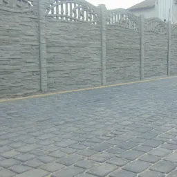 Długi, szary mur z prefabrykowanych elementów betonowych imitujących kamień, zwieńczony dekoracyjnym ażurem, oddzielający posesję od wybrukowanej drogi z szarej kostki.