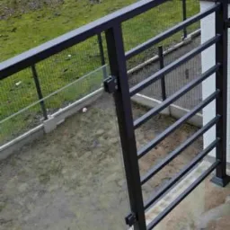 Ciemna, metalowa balustrada balkonowa z prostymi, poziomymi szczeblami i kwadratowymi słupkami, widok z góry na zewnątrz, z zielonym trawnikiem w tle.