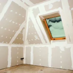 Poddasze w trakcie wykańczania płytami gipsowo-kartonowymi, widoczne łączenia wzmocnione taśmą i szpachlowane, okno dachowe w drewnianej ramie, surowy beton na podłodze.