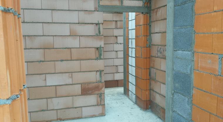 Wnętrze budynku w trakcie budowy, ściany z cegły ceramicznej i bloczków betonowych, widoczne spoiny zaprawy, przygotowane otwory drzwiowe.