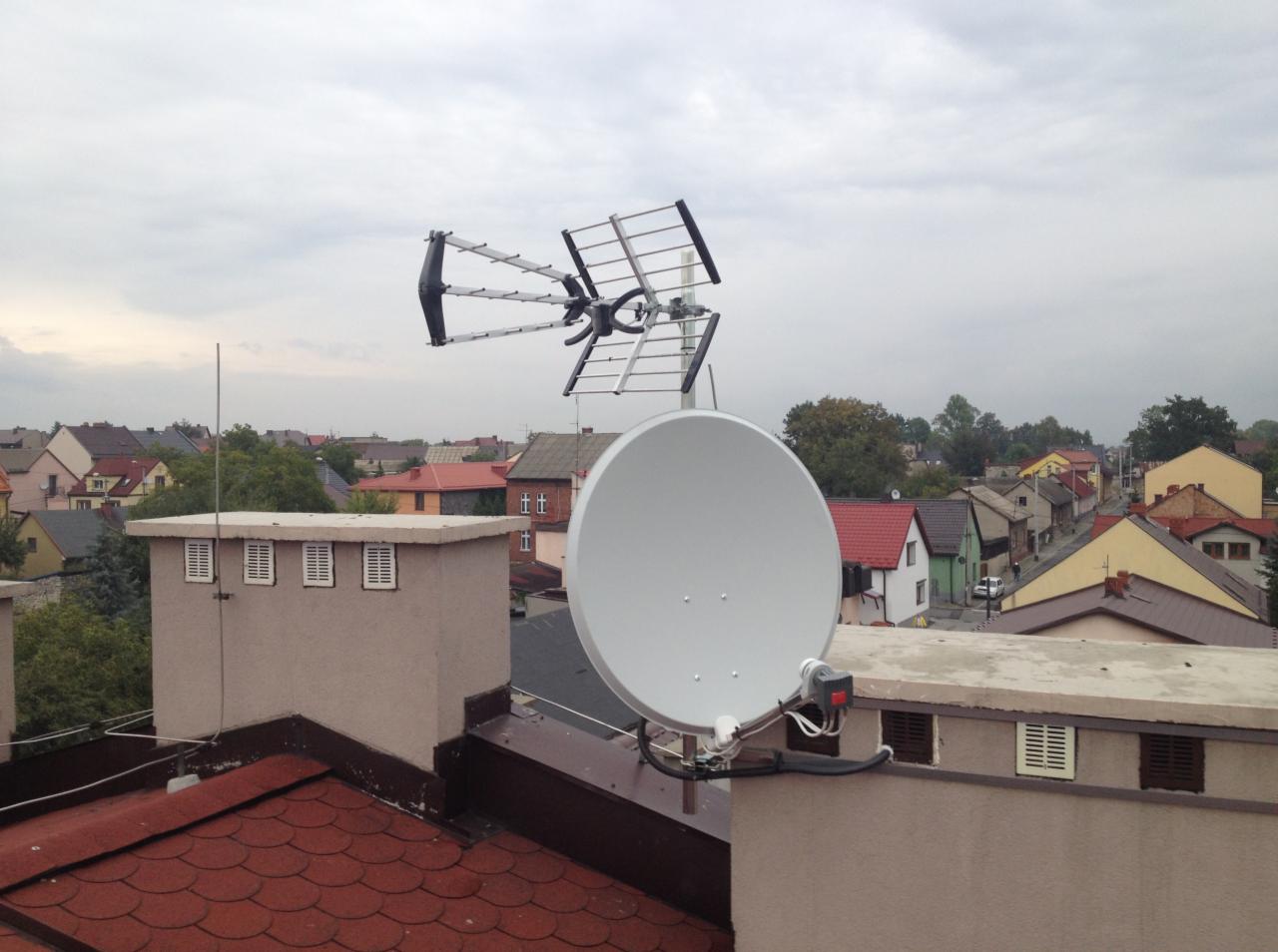 Antena satelitarna i antena naziemna zamontowane na dachu budynku mieszkalnego z widokiem na okoliczną zabudowę w pochmurny dzień.
