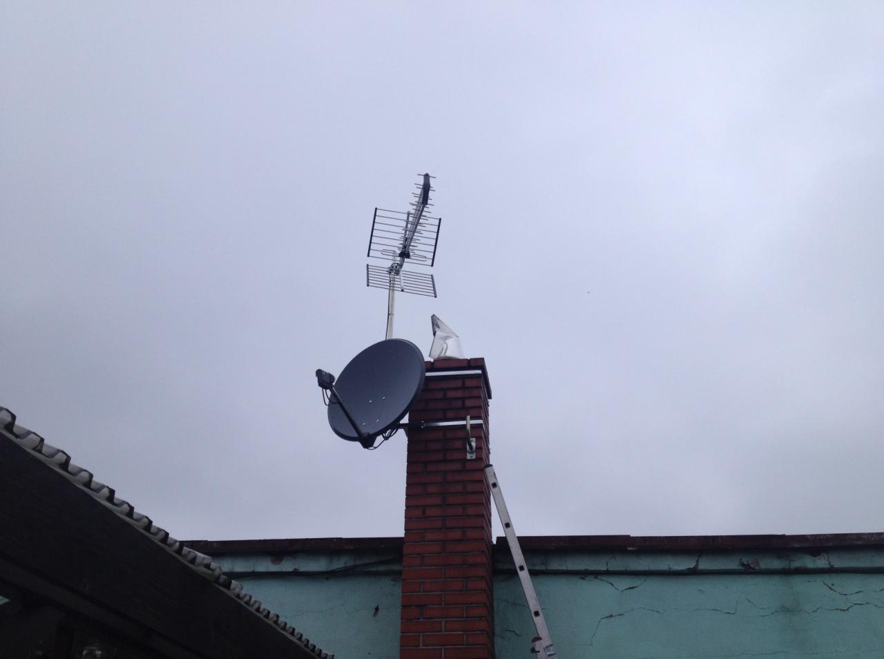 Antena satelitarna i antena naziemna zamontowane na kominie z drabina opartą o ścianę budynku w pochmurny dzień.