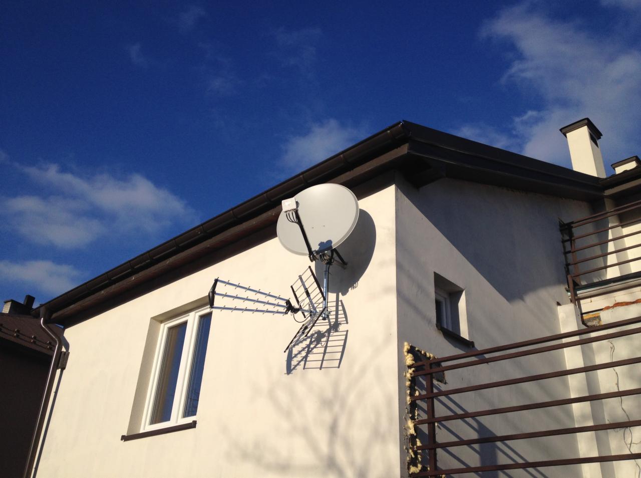 Biały dom z zamontowaną anteną satelitarną i anteną naziemną, widoczne ciemne rynny i błękitne niebo z chmurami.
