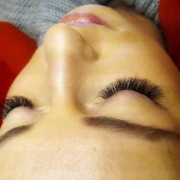 Gabinet Kosmetyki Profesjonalnej Lumiere Julita Stępień - Zbliżenie na twarz kobiety z przedłużonymi, gęstymi rzęsami, widoczne brwi, delikatny błyszczyk na ustach, czerwona poduszka w tle.