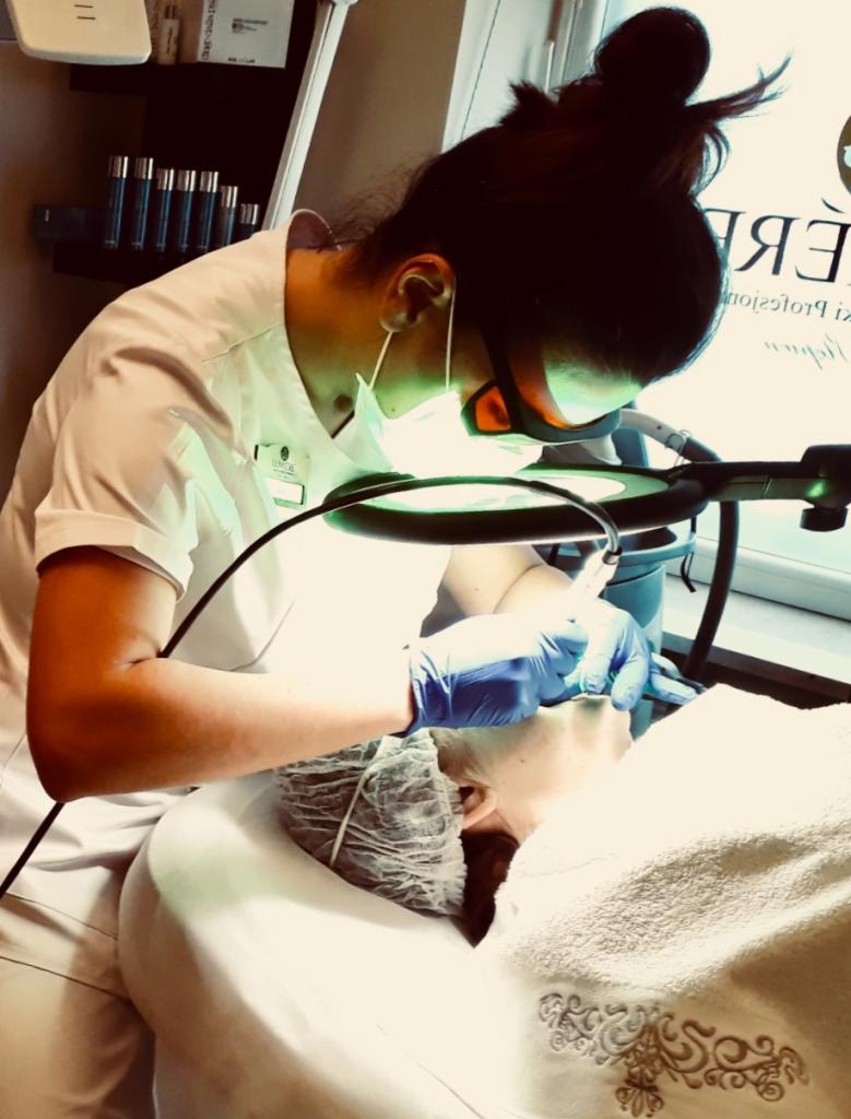 Osoba w uniformie i ochronnych okularach wykonuje zabieg na twarzy klientki leżącej na leżance, oświetlonej lampą powiększającą.