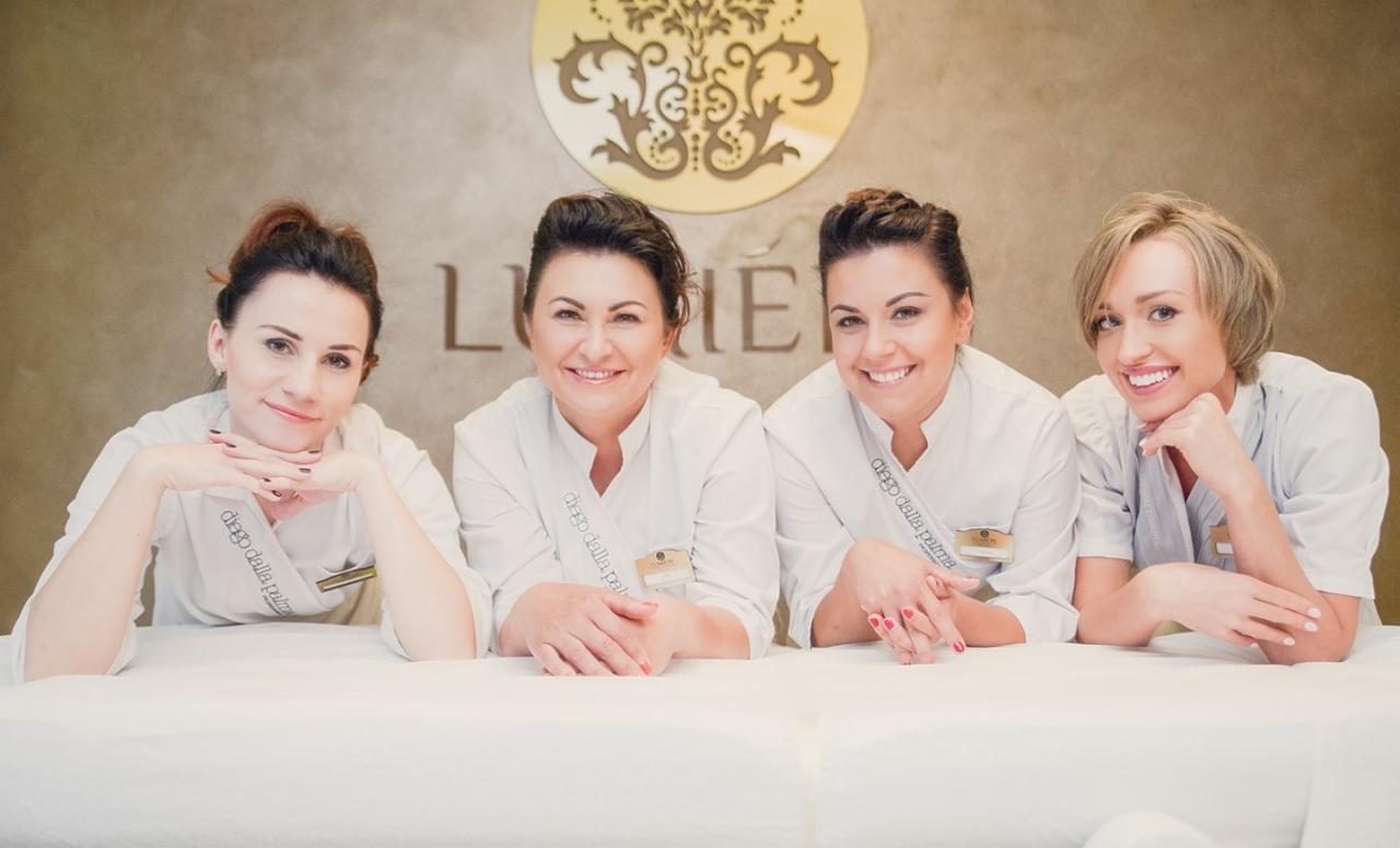 Uśmiechnięty zespół czterech kosmetyczek w białych uniformach, z logo firmy na ścianie w tle, opierający się o biały stół lub leżankę.