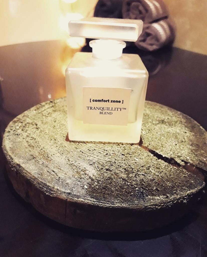 Kwadratowa butelka z matowego szkła z napisem '[comfort zone] TRANQUILLITY™ BLEND' na drewnianym krążku z widocznym pęknięciem i szorstką powierzchnią.