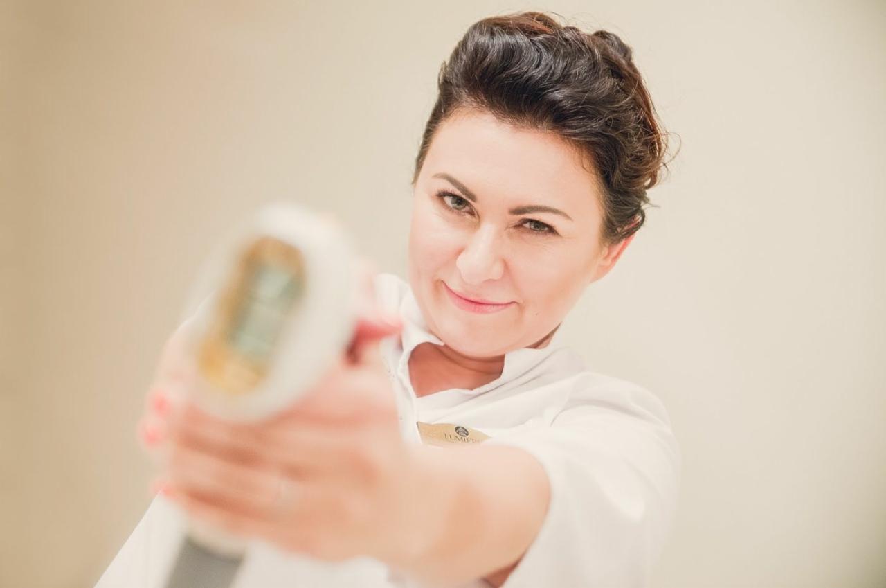 Kobieta w białym uniformie celuje urządzeniem do depilacji laserowej, patrząc wprost w obiektyw z lekkim uśmiechem.