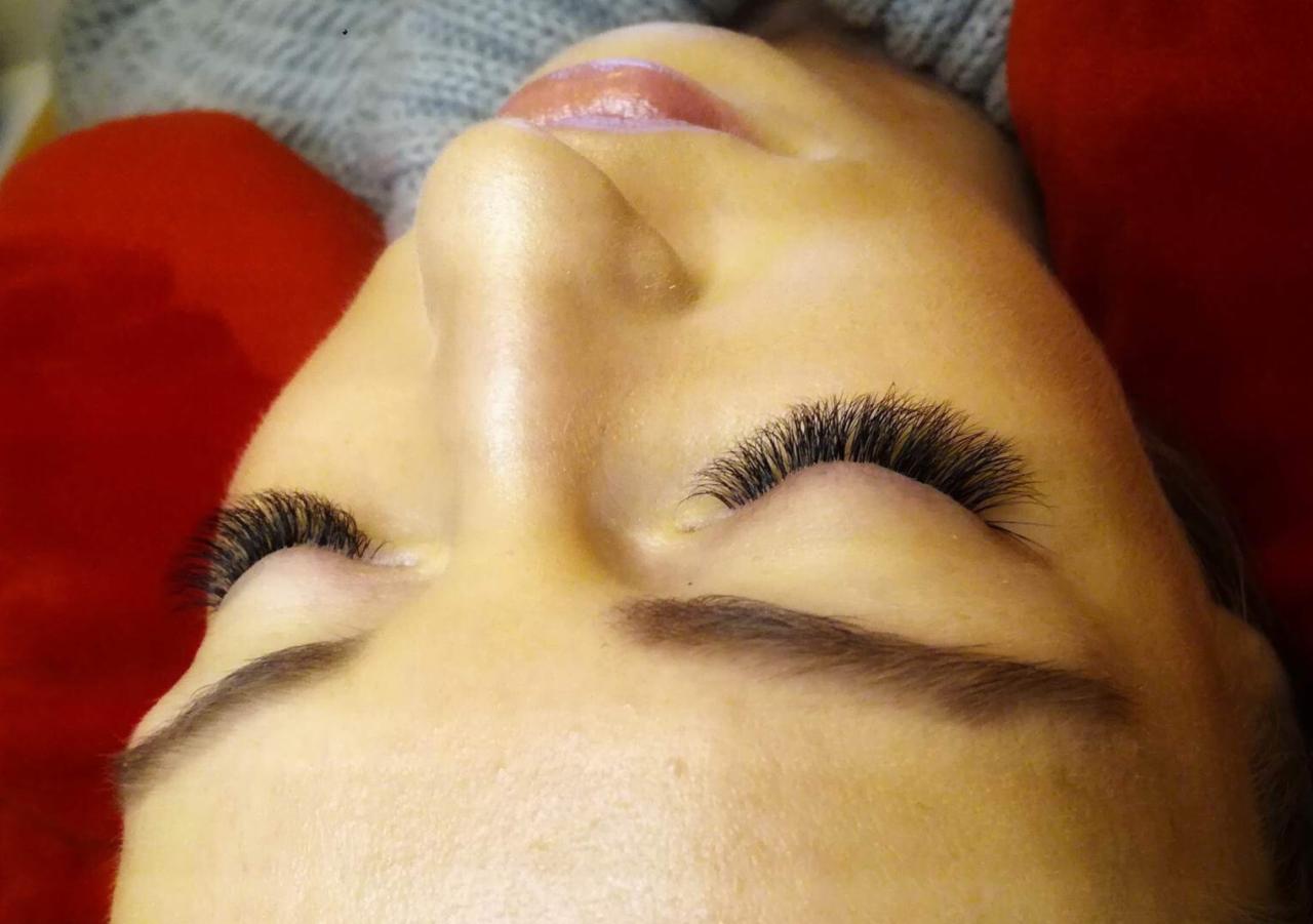 Zbliżenie na twarz kobiety z przedłużonymi, gęstymi rzęsami, widoczne brwi, delikatny błyszczyk na ustach, czerwona poduszka w tle.