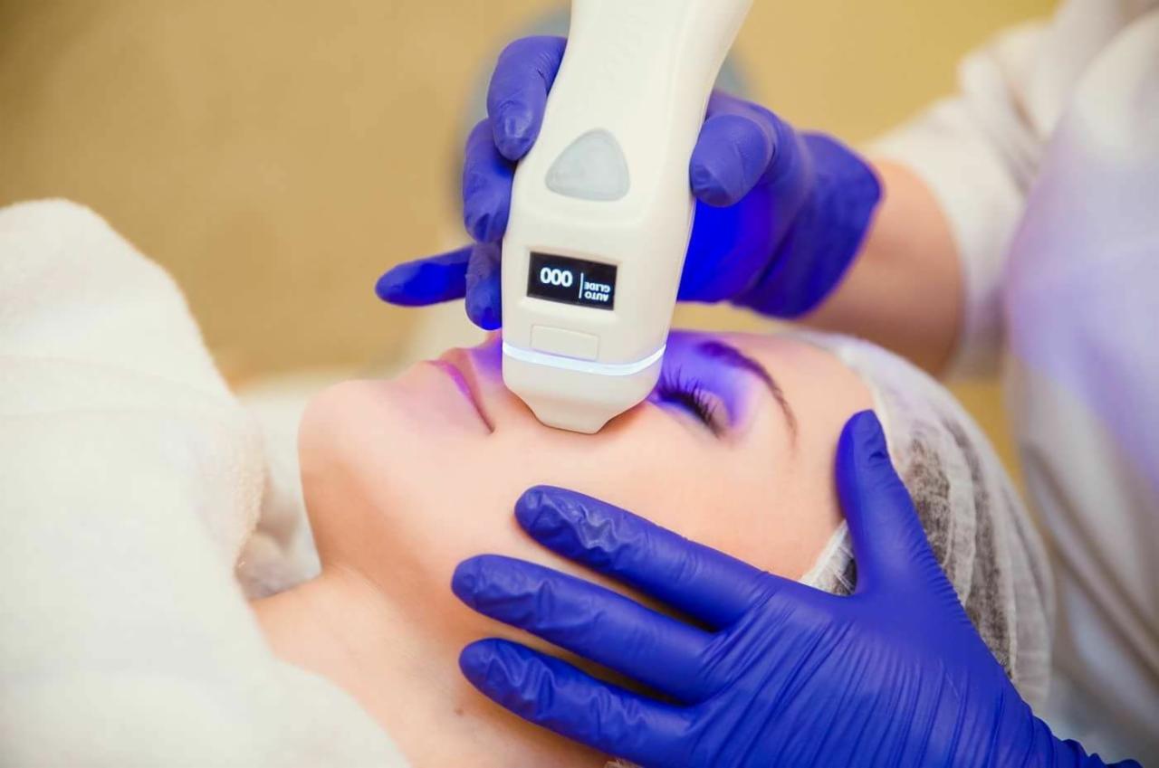 Zabieg na twarz z użyciem urządzenia emitującego niebieskie światło, wykonywany przez osobę w niebieskich rękawiczkach ochronnych; pacjentka w czepku kosmetycznym leży na leżance.