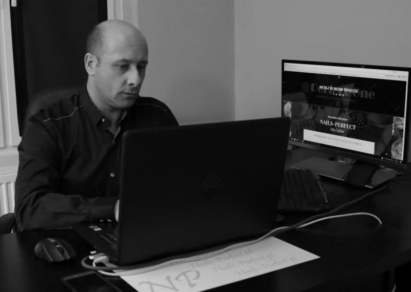 Mężczyzna skupiony na pracy przy laptopie, na ekranie monitora widoczna strona internetowa 'Nails-Perfect', obok notatka z adresem strony, czarno-biała fotografia.