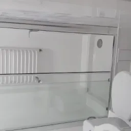 Biała łazienka z kabiną prysznicową ze szklanymi drzwiami przesuwnymi, grzejnikiem drabinkowym i toaletą z kompaktową spłuczką, wykończona płytkami imitującymi marmur.