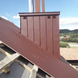 Montaż brązowego, falistego pokrycia dachowego z blachy na drewnianej konstrukcji więźby, widoczny komin z blachy w tym samym kolorze, w tle zabudowania i wzgórza.