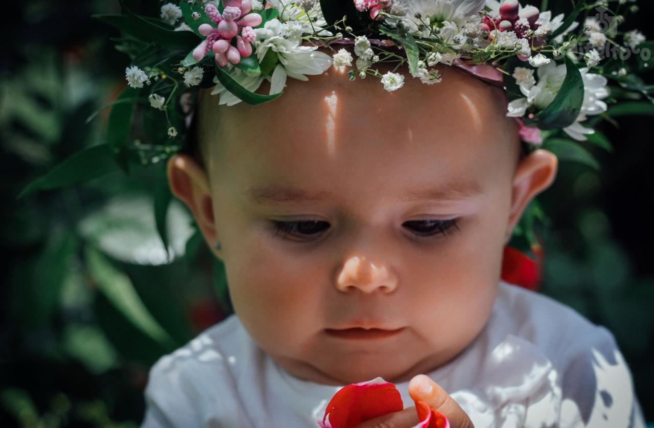 Portret niemowlęcia z wiankiem z białych i różowych kwiatów na głowie, trzymającego w dłoni płatek róży.