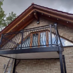 Zewnętrzny balkon z czarną, metalową balustradą o zdobnym wzorze, zamontowany na ceglanym budynku z drewnianym dachem i oświetleniem zewnętrznym.