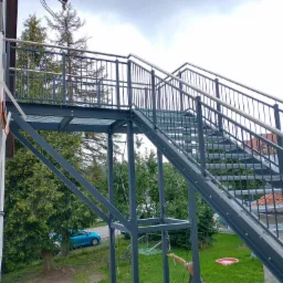 Zewnętrzne schody metalowe z balustradami, prowadzące na piętro budynku, konstrukcja pomalowana na szaro, widok z dołu na tle zieleni i nieba.