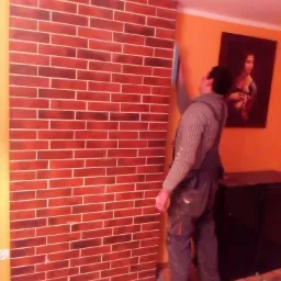 Pracownik w kombinezonie wygładza ścianę z efektem cegły w pomarańczowym pokoju z obrazem Mona Lisy w tle.