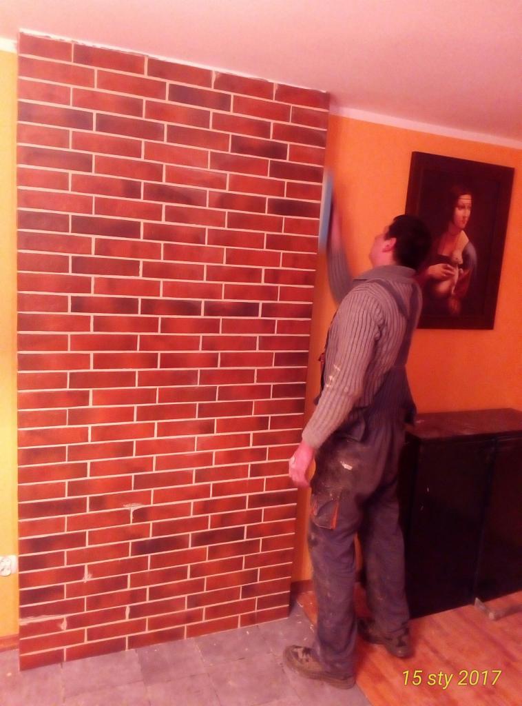 Pracownik w kombinezonie wygładza ścianę z efektem cegły w pomarańczowym pokoju z obrazem Mona Lisy w tle.