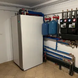 Biała jednostka Nibe VVM 500 z zieloną diodą, podłączona do niebieskich i czarnych rur, oraz dwa niebieskie zbiorniki ciśnieniowe i czarne moduły sterujące z manometrami, zamontowana...