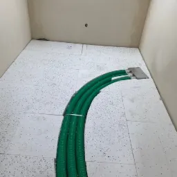 Trzy zielone, karbowane rury wentylacyjne, spięte metalową opaską, biegną po białej podłodze z czarnymi kropkami w surowym pomieszczeniu z beżowymi ścianami.