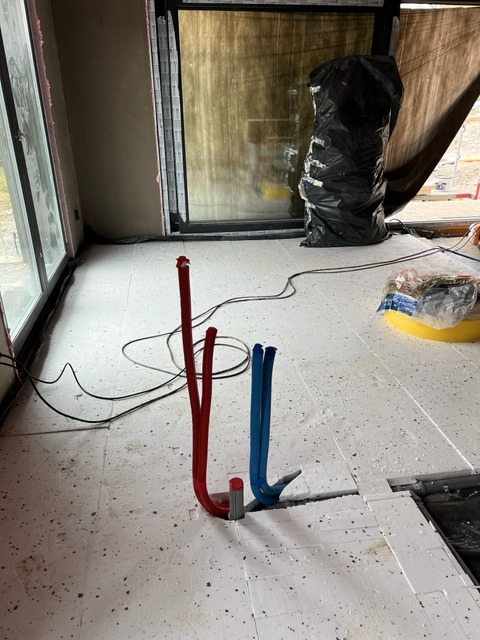 Rury grzewcze czerwone i niebieskie wystające z białej izolacji podłogowej, widoczne okna i czarny worek na śmieci w tle.