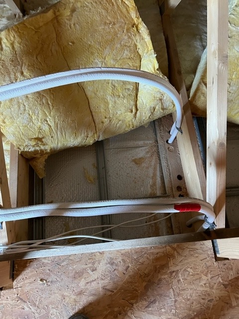 Fragment instalacji z izolowanymi rurami w przestrzeni poddasza z widoczną wełną mineralną i elementami konstrukcji drewnianej.