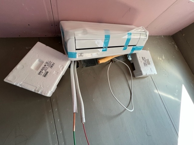 Klimatyzator typu split, jeszcze w folii ochronnej z niebieską taśmą, przygotowany do montażu, widoczne przewody elektryczne i rurki chłodnicze, obok styropianowe elementy opakowania.