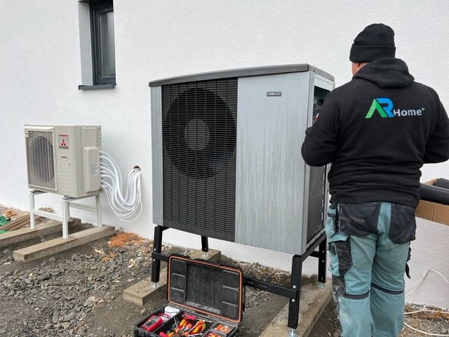 Instalacja zewnętrznej jednostki systemu grzewczego, widoczna skrzynka z narzędziami i pracownik w bluzie z logo firmy, rury doprowadzające do drugiej jednostki na wspornikach.