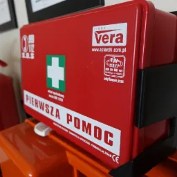 Czerwona apteczka pierwszej pomocy firmy Vera zamocowana na ścianie, obok gaśnicy.