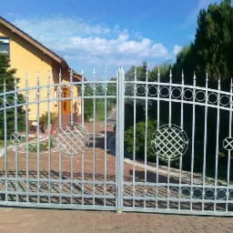 Dwuskrzydłowa metalowa brama wjazdowa z ozdobnymi elementami w kształcie kół i kwadratów na tle domu i zieleni.