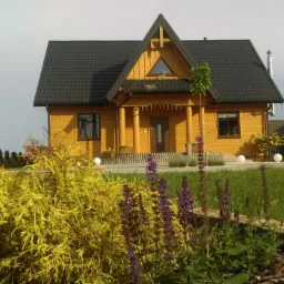 Żółty drewniany dom z czarnym dachem, kolumnami przy wejściu i elementami dekoracyjnymi, otoczony zielonym trawnikiem, krzewami i kwiatami, z kulistymi lampami ogrodowymi w tle.