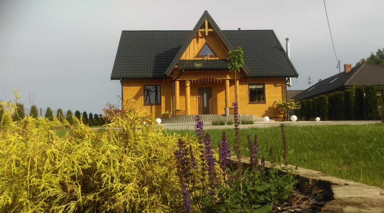 Żółty drewniany dom z czarnym dachem, kolumnami przy wejściu i elementami dekoracyjnymi, otoczony zielonym trawnikiem, krzewami i kwiatami, z kulistymi lampami ogrodowymi w tle.