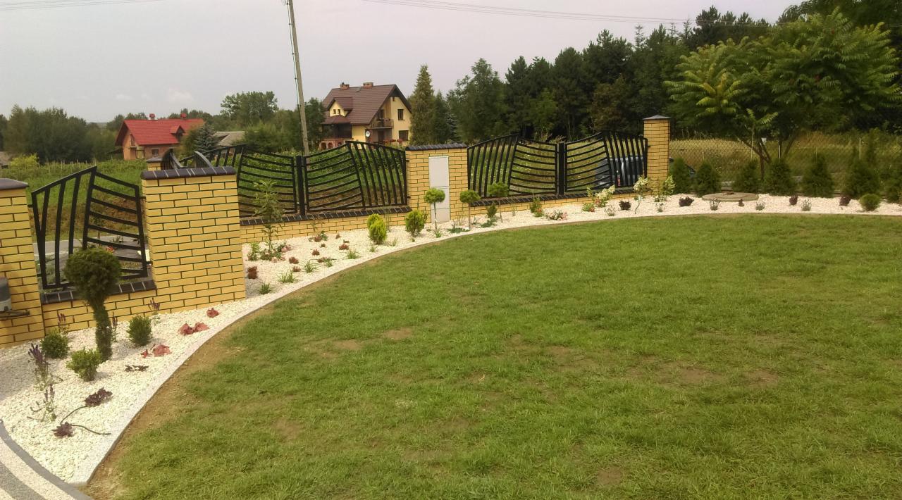 Uporządkowany ogród z równo przystrzyżonym trawnikiem, obrzeżem z białego kamienia i niskim, ozdobnym ogrodzeniem z cegły i metalu, w tle domy i drzewa.