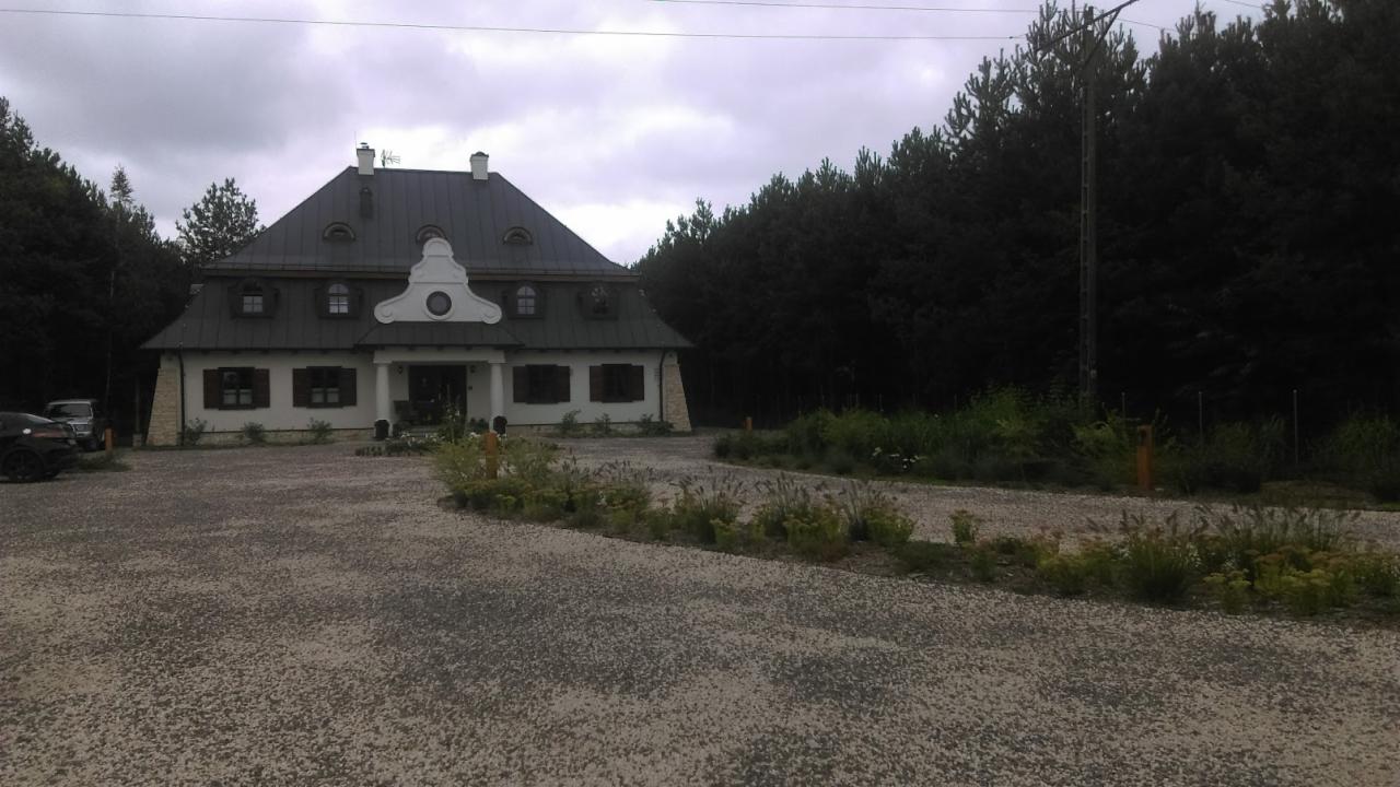 Dwór w stylu dworkowym z ciemnym dachem, białymi ścianami i ciemnymi okiennicami, otoczony lasem i żwirowym podjazdem z niską zielenią.
