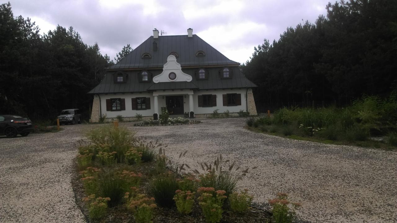 Dwupiętrowy dom z ciemną dachówką i białymi ścianami, otoczony lasem i żwirowym podjazdem z rabatami kwiatowymi.