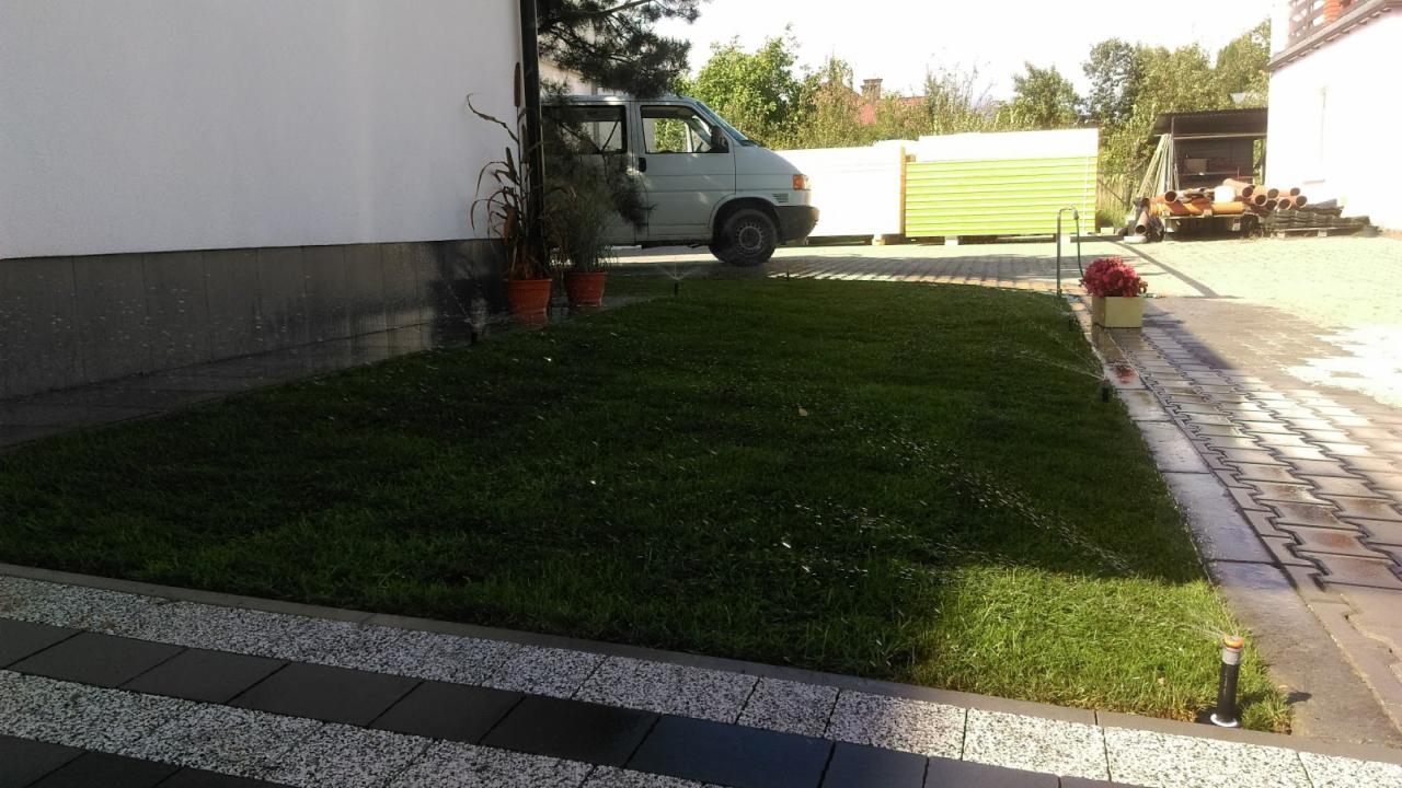 Świeżo ułożona trawa z rolki, nawadniana przez system zraszaczy, z zaparkowanym białym vanem w tle i widocznymi donicami z roślinami oraz elementami budowlanymi.